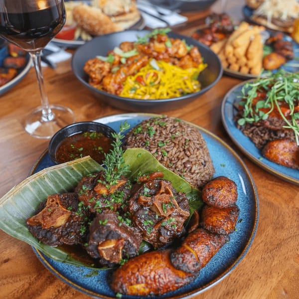 A blue plate of Bacchanal Junction's oxtail, rice and peas, plantains, and dipping sauce, with other dishes blurred in the background.
