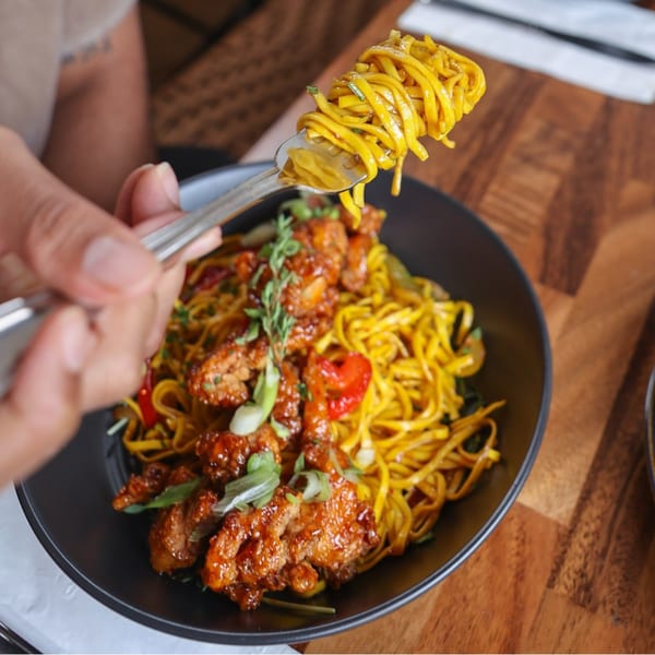A fork lifts bright yellow noodles from a black bowl of Caribbean chicken and noodles.