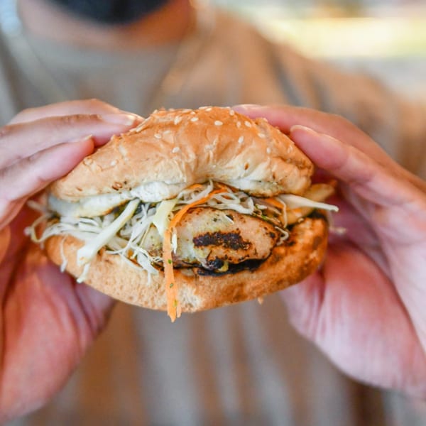 Hands holding a Bacchanal Junction Caribbean jerk chicken sandwich with coleslaw on a sesame bun.
