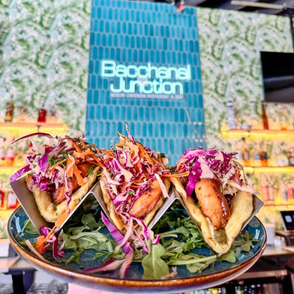 Three Caribbean tacos with shrimp, red cabbage, and carrots on a blue plate with Bacchanal Junction neon sign.